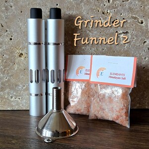 May include: Two silver metal grinders with a black top and a clear window on the side. A silver metal funnel is in front of the grinders. Two small bags of pink Himalayan salt are in the background. The bags have the text "ELEMIANTS Himalayan Salt" printed on them.