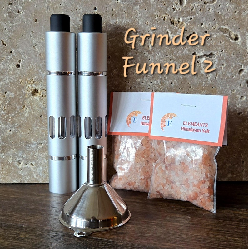 May include: Two silver metal grinders with a black top and a clear window on the side. A silver metal funnel is in front of the grinders. Two small bags of pink Himalayan salt are in the background. The bags have the text "ELEMIANTS Himalayan Salt" printed on them.