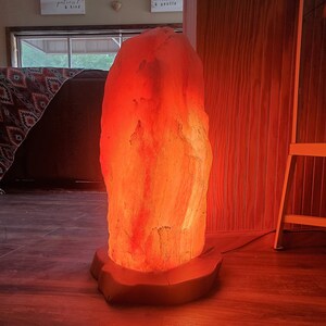 Pode incluir: Uma grande l&acirc;mpada de sal do Himalaia de cor laranja com uma base de madeira. A l&acirc;mpada est&aacute; acesa e brilha, projetando uma luz laranja quente.