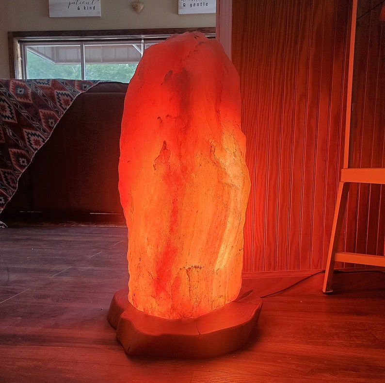 Pode incluir: Uma grande l&acirc;mpada de sal do Himalaia de cor laranja com uma base de madeira. A l&acirc;mpada est&aacute; acesa e brilha, projetando uma luz laranja quente.