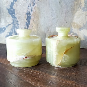 May include: Two matching, cylindrical, lidded jars made of translucent, pale green onyx. The jars have a stepped base and a rounded knob on the lid. The onyx has natural veining with streaks of white, brown, and red.