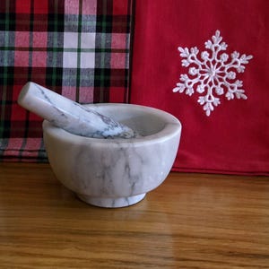 May include: A white marble mortar and pestle set. The mortar is round and has a white marble base. The pestle is long and has a white marble handle.