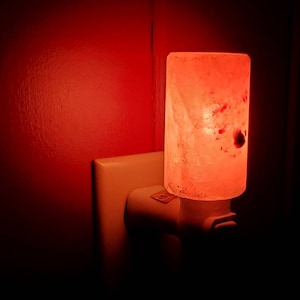 May include: A rectangular Himalayan salt night light plugged into a wall outlet. The salt lamp glows with a warm, reddish-orange light. The outlet and surrounding wall are a light beige and red, respectively.