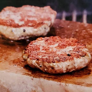 May include: Two round, cooked hamburger patties on a pink Himalayan salt block. The patties are browned and have a slightly crispy texture.
