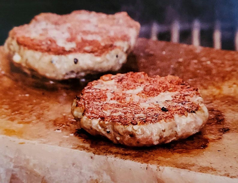 May include: Two round, cooked hamburger patties on a pink Himalayan salt block. The patties are browned and have a slightly crispy texture.