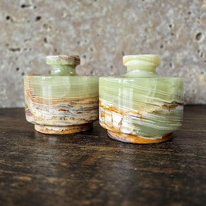 May include: Two small, decorative onyx jars with lids. The jars are cylindrical with a light green hue and brown and white veining. The lids are a lighter green. The jars sit on a dark wooden surface.