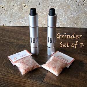May include: Two silver salt grinders with black tops and clear windows, labeled "Grinder Set of 2". Two small bags of pink Himalayan salt are included.