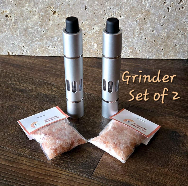 May include: Two silver salt grinders with black tops and clear windows, labeled "Grinder Set of 2". Two small bags of pink Himalayan salt are included.