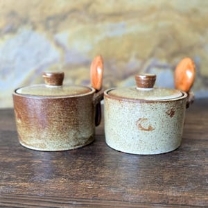 May include: Two ceramic lidded jars with attached wooden spoons. The jars are brown and beige with a speckled texture. The lids have small, rounded knobs. The wooden spoons are a light orange color.