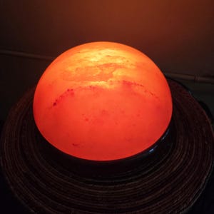 Może przedstawiać: Duża, okrągła, pomarańczowa lampa solna z Himalaj&oacute;w z czarną podstawą. Lampa jest włączona i świeci.
