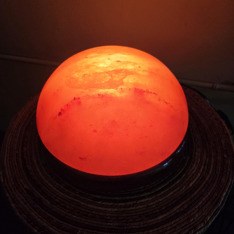 Może przedstawiać: Duża, okrągła, pomarańczowa lampa solna z Himalaj&oacute;w z czarną podstawą. Lampa jest włączona i świeci.