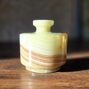 May include: A small, decorative onyx jar with a lid. The jar is primarily light green with bands of brown and tan. The lid has a small handle. The jar is likely used for storing small items or as a decorative piece.