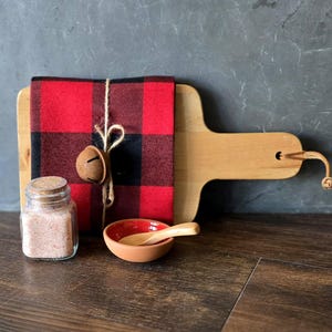 May include: A red and black checkered kitchen towel tied with twine and a small bell, a wooden cutting board with a leather handle, a small glass jar of pink Himalayan salt, and a small red ceramic bowl with a wooden spoon.