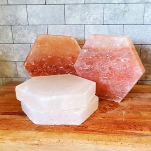 May include: Three hexagonal salt lamps, two are pink Himalayan salt and one is white Himalayan salt. The salt lamps are on a wooden surface.