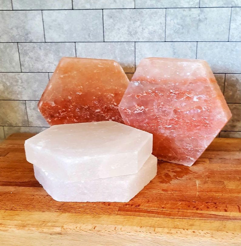 May include: Three hexagonal salt lamps, two are pink Himalayan salt and one is white Himalayan salt. The salt lamps are on a wooden surface.
