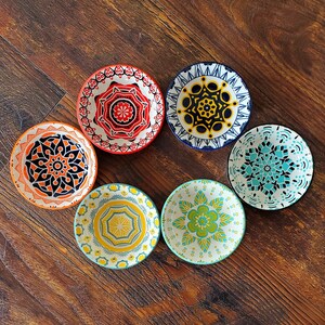 May include: Six colorful ceramic bowls with intricate geometric patterns. The bowls are in shades of blue, red, yellow, and white. The bowls are arranged in a circle on a wooden surface.