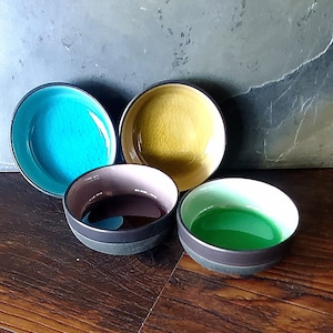 May include: Four small ceramic bowls with different colored glazes. The bowls are arranged in a semi-circle on a wooden surface. The bowls are turquoise, yellow, brown, and green.