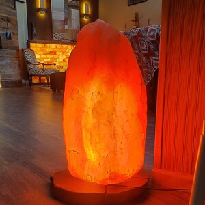 Pode incluir: Uma grande l&acirc;mpada de sal do Himalaia laranja com uma base de madeira. A l&acirc;mpada est&aacute; acesa e brilhando, projetando uma luz laranja quente.