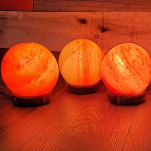 May include: Three spherical Himalayan salt lamps, each emitting a warm orange glow. The lamps sit on dark bases, with visible marbling patterns on their surfaces. The background features a wooden wall and floor.