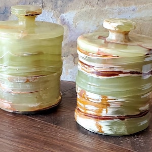 May include: Two matching decorative jars with lids. The jars are cylindrical with a light green and brown banded pattern. The lids have a small knob on top. The jars are made of a translucent stone material.
