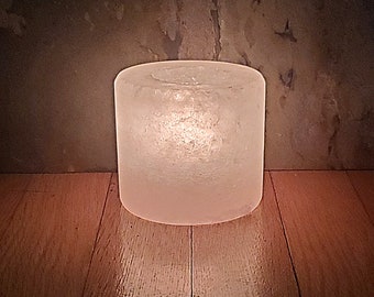 Könnte beinhalten: Ein gefrosteter Glas-Kerzenhalter mit runder Form und glatter, weißer Oberfläche. Der Kerzenhalter ist angezündet und strahlt ein warmes Licht aus.