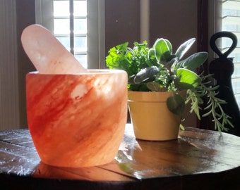 May include: A pink Himalayan salt mortar and pestle set. The mortar has a rounded shape with a swirling pattern of pink and white. A small yellow pot with green herbs sits to the right.