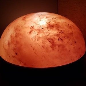 Może przedstawiać: Okrągła, r&oacute;żowa lampa solna z Himalaj&oacute;w z ciepłym, pomarańczowym blaskiem. Lampa wykonana jest z naturalnych kryształ&oacute;w soli i ma fakturowaną powierzchnię.