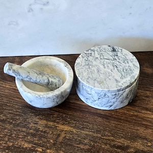 May include: A marble mortar and pestle set with a matching round lidded container. The set features a white and gray veined pattern. The mortar is round with a pestle resting inside. The container is also round.