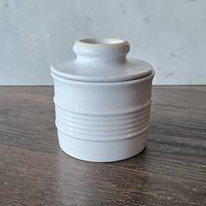 May include: A white ceramic butter dish with a lid. The dish has a cylindrical base with horizontal ridged detailing. The lid has a small knob and a slightly off-white rim. The butter dish is designed to keep butter fresh.