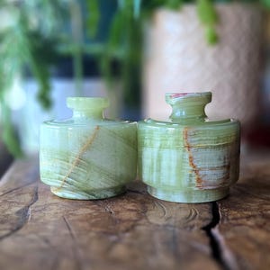 May include: Two green onyx decorative jars with a cylindrical body and a small, rounded top. The jars have natural veining in shades of green, brown, and white. They are displayed on a wooden surface.