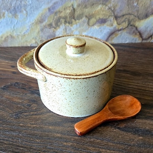 May include: A speckled cream-colored ceramic sugar bowl with a matching lid and a single side handle. The bowl features a small, round knob on the lid and is accompanied by a small, smooth, polished wooden spoon with a rounded bowl shape.