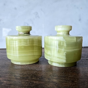 May include: Two small, cylindrical containers crafted from light green banded stone. Each vessel features a smooth, polished finish with visible horizontal layers of translucent mineral patterns, a short neck, and a flat, circular base.
