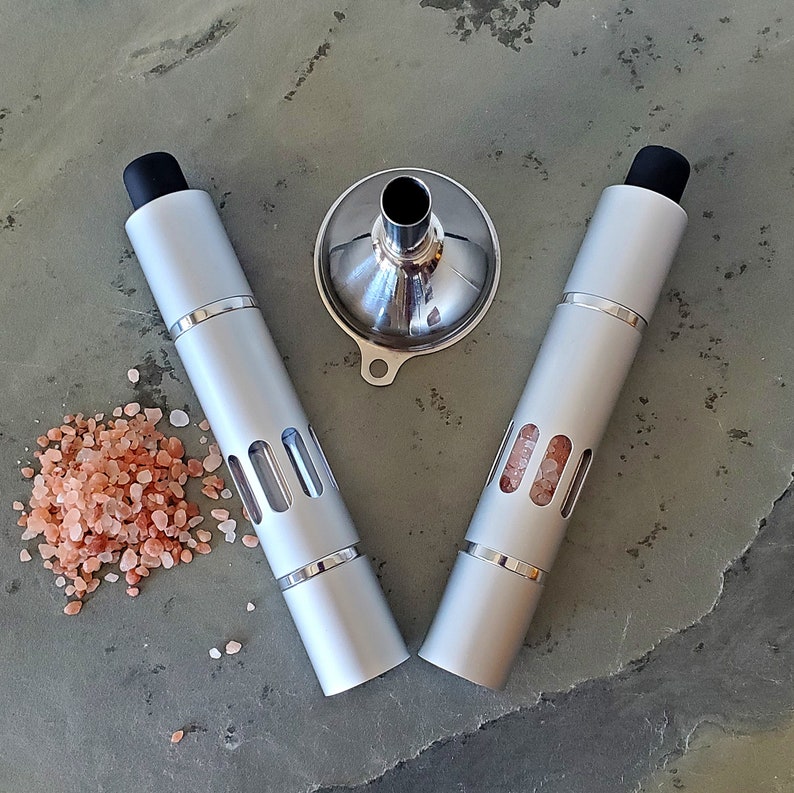 May include: Two silver metal salt grinders with black tops and a silver metal funnel. The grinders have clear windows to see the salt inside. Pink Himalayan salt is scattered around the grinders.