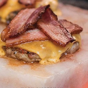 May include: A cheeseburger topped with crispy bacon is cooked on a Himalayan salt block. The salt block is pink and has a rough texture. The burger is cooked to a golden brown and the cheese is melted and bubbly.