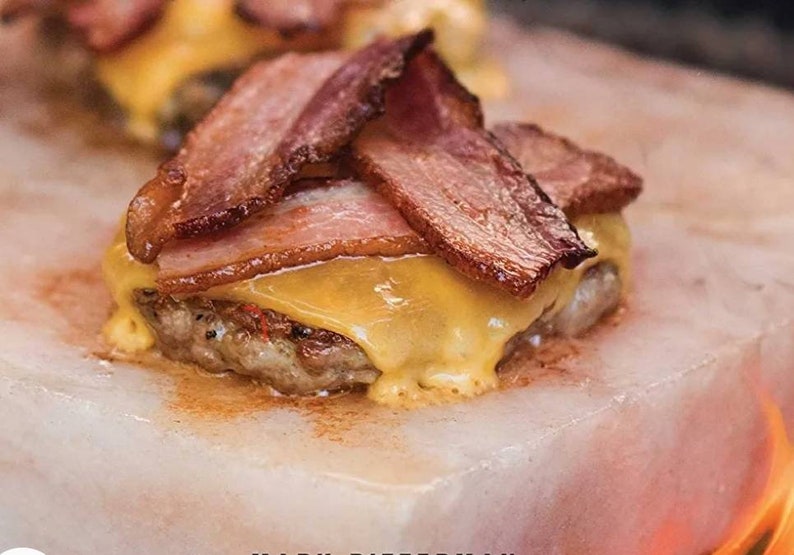 May include: A cheeseburger topped with crispy bacon is cooked on a Himalayan salt block. The salt block is pink and has a rough texture. The burger is cooked to a golden brown and the cheese is melted and bubbly.
