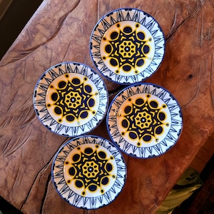 May include: Four small, round decorative plates with a white background, blue rims, and a central design of yellow and navy blue geometric shapes. The plates are arranged on a wooden surface.