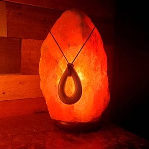 May include: A Himalayan salt lamp with a warm orange glow, featuring a decorative pendant hanging from the top. The pendant is a dark brown, oval-shaped piece with a hole in the center.