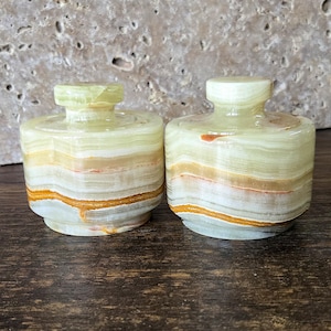 May include: Two decorative onyx jars with lids. The jars are cylindrical with a rounded top and base. The onyx has layers of light green, white, and brown. The jars are on a dark wooden surface.