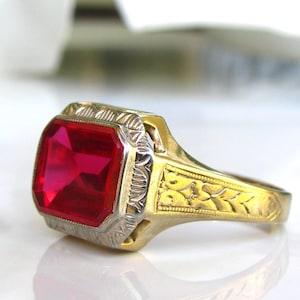May include: A gold ring with a large, red, square-shaped gemstone set in a bezel setting. The band is decorated with a floral design.