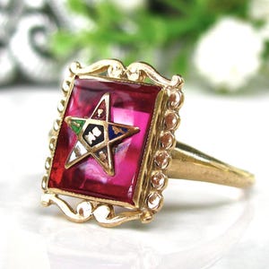 May include: A gold-toned ring with a square, ruby-red center. The center features a white star with colorful accents and a gold border. The ring's design includes ornate detailing around the edges.