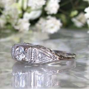 May include: A silver ring with a large, round, clear diamond in the center. The ring has a delicate, intricate design with smaller diamonds on either side of the center stone.