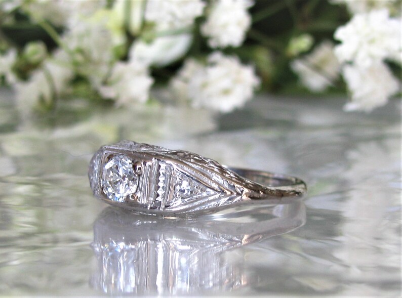 May include: A silver ring with a large, round, clear diamond in the center. The ring has a delicate, intricate design with smaller diamonds on either side of the center stone.