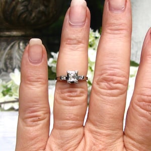 May include: A silver engagement ring with a square-shaped diamond set in a prong setting. The ring is on a finger with a white background.