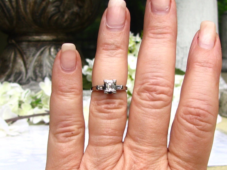 May include: A silver engagement ring with a square-shaped diamond set in a prong setting. The ring is on a finger with a white background.