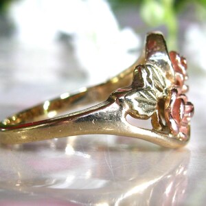 May include: A gold-tone ring featuring a floral design with two roses and leaves. The roses have a reddish-pink hue, and the ring's band is smooth and polished. The ring is likely made of gold or a gold-plated metal.