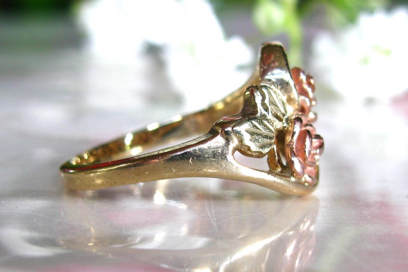 May include: A gold-tone ring featuring a floral design with two roses and leaves. The roses have a reddish-pink hue, and the ring's band is smooth and polished. The ring is likely made of gold or a gold-plated metal.