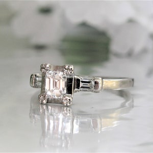 May include: A silver engagement ring with a square-shaped diamond and two smaller baguette diamonds on the band.