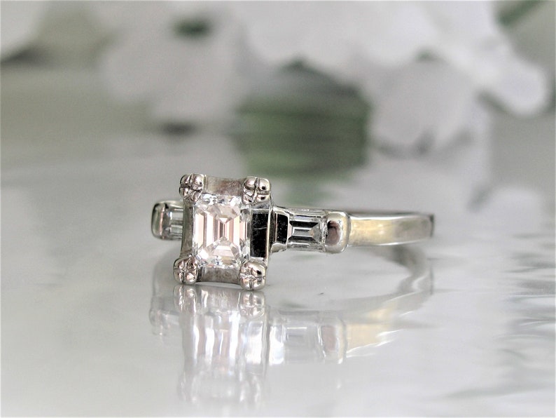 May include: A silver engagement ring with a square-shaped diamond and two smaller baguette diamonds on the band.