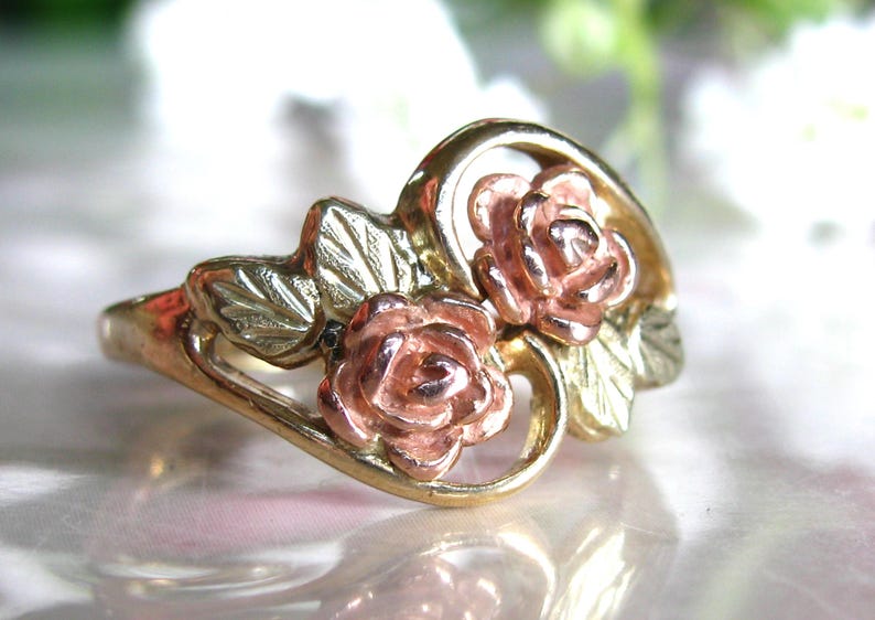 May include: A gold-tone ring featuring two rose blossoms in a pink hue, with gold and silver-tone leaves. The ring has a delicate, floral design, perfect for a romantic or nature-inspired aesthetic.