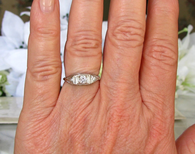 May include: A silver ring with a single round diamond and smaller diamonds on each side. The ring is on a person's finger.